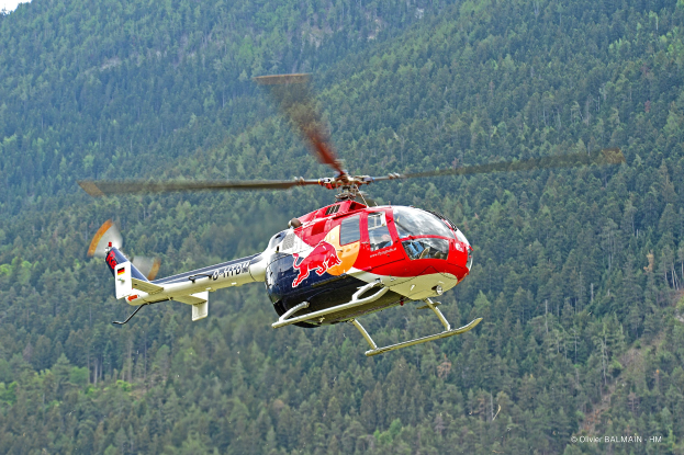 Roter Bullen-Hubschrauber über einem grünen Wald mit Bäumen auf den Höhen im Hintergrund und einem klaren blauen Himmel öber.