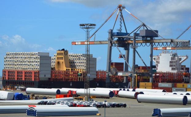 Ein großes Container-Schiff liegt an einem Hafen mit verschiedenen Geräten und Gebäuden im Hintergrund unter einem bewölkten Himmel.