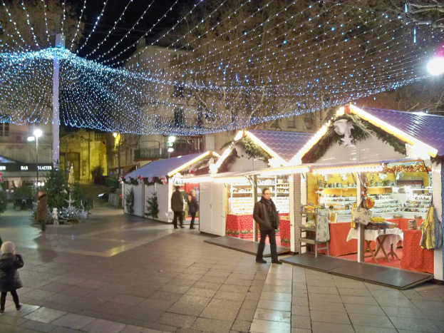 Ein nächtlicher Weihnachtsmarkt in einer Stadt, mit Menschen, die stehen, sitzen und eine Person ein Kind hält, umgeben von festlichen Lichtern, Pfählen, Pflanzen, Bäumen und Gebäuden mit Fenstern unter einem bewölkten Himmel.