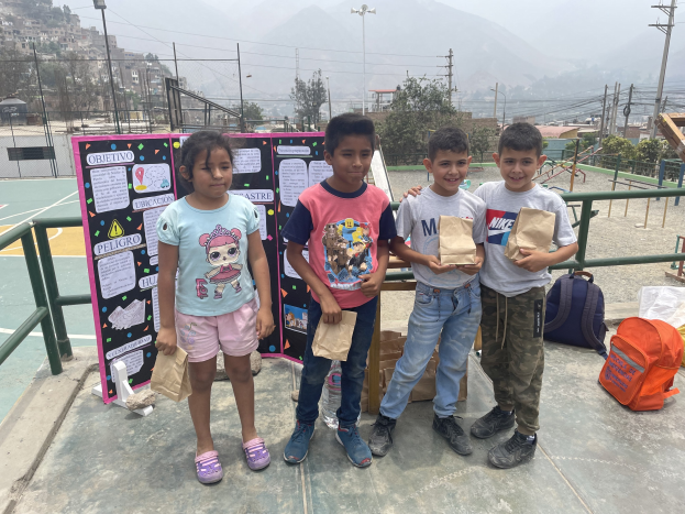 Gruppe von Kindern auf einem Basketballfeld mit Papierbeuteln in der Hand, mit Gegenständen auf dem Boden rechts und einer Tafel mit Text links, vor einem Hintergrund aus Zaun, Pfosten, Straßenlaternen, Bäumen, Gebäuden, Hügeln und Himmel.