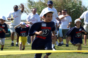 Kinder laufen bei einem Wettbewerb auf einem grünen Rasenfeld mit einem gelben Band im Vordergrund, einige tragen Kappen und Schwimmbrillen, andere halten Gegenstände, vor einem Hintergrund aus Bäumen und einem klaren blauen Himmel.