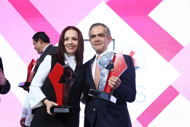 Zwei Personen auf einer Bühne, die lächeln und Trophäen halten, mit einem sichtbaren Bildschirm dahinter.