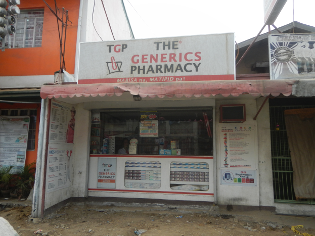 Eine Apotheke namens "The Generics Pharmacy" umgeben von Gebäuden, Strommasten, Stromkabeln, Werbetafeln, Topfpflanzen, Kraftfahrzeugen auf der Straße, unter einem klaren blauen Himmel.