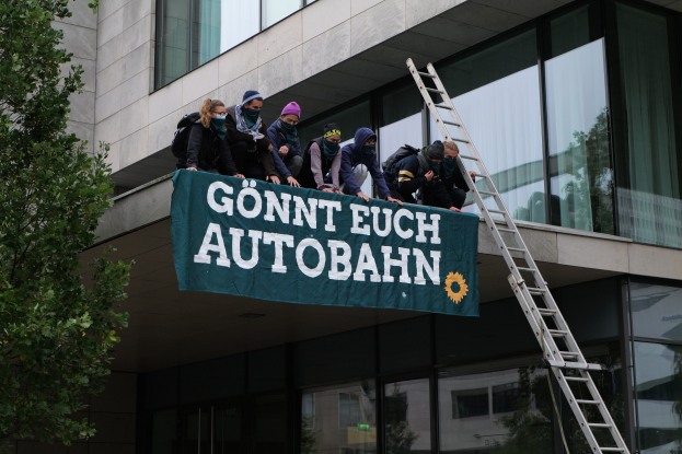 Eine Gruppe von Menschen auf einem Gebäude mit einer Fahne, mit einem Baum links und Glasfenstern im Hintergrund, die scheinbar protestieren und die deutsche Regierung unterstützen.