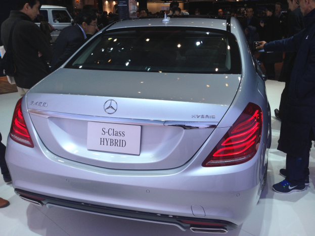 Silberner Mercedes S-Klasse Hybrid auf einer Automesse präsentiert, umgeben von Menschen in verschiedenenfarbigen Kleidern, mit anderen Fahrzeugen und Beleuchtung im Hintergrund.