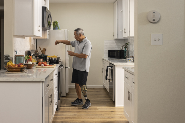 Ein Mann steht in einer Küche mit weißen Schränken und hält ein Glas in der Hand, während verschiedene Gegenstände wie Früchte, Gemüse und Utensilien auf einer Arbeitsplatte links von ihm liegen.