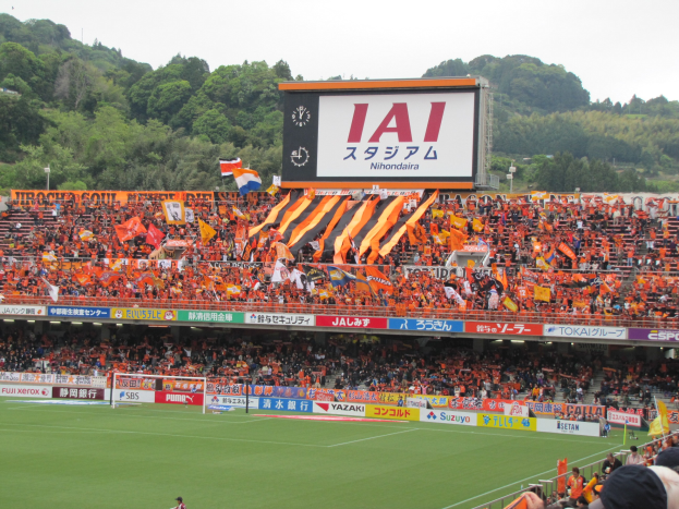 Fußballspiel in einem Stadion mit großer Zuschauermenge, grünem Rasen, Torstangen, Bannern, Fahnen, einem großen Bildschirm, Bäumen und einem klaren blauen Himmel.