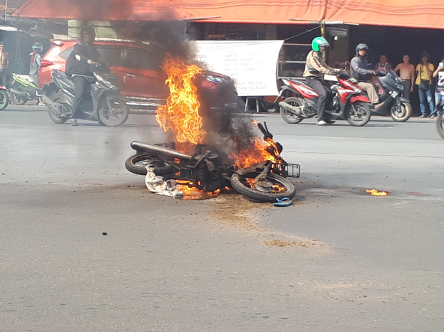 Motorrad in Flammen an der Straße mit einem Auto und einem Van im Hintergrund, umgeben von Menschen, einige mit Helmen, eine Fahne mit Text und ein Zelt.