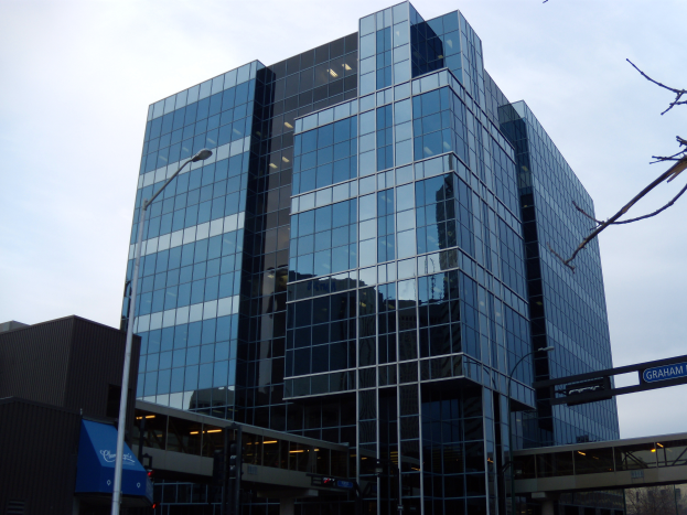 Ein hohes Glasgebäude mit vielen Fenstern in einer Stadt, umgeben von Straßenmasten, Straßenlaternen, Namensschildern, Verkehrspfosten, Verkehrssignalen, Kraftfahrzeugen, Bäumen und einem bewölkten Himmel.