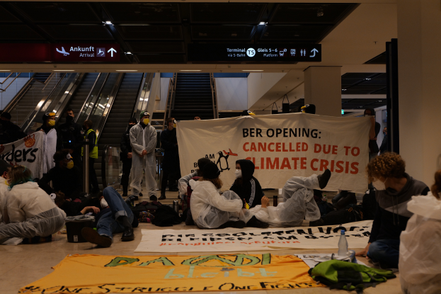 Eine Gruppe von Menschen sitzt auf dem Boden eines Flughafens und hält ein Banner in der Hand, auf dem "Berlin wegen Klimakrise gestrichen" steht. Um sie herum liegen Taschen, Flaschen und andere Gegenstände verstreut, im Hintergrund sind Rolltreppen, Tafeln mit Text und Deckenleuchten zu sehen.