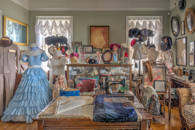Ein Raum im Kunstmuseum in St. Louis, Missouri, voller Schaufensterpuppen in Kleidern und H├Ątchen, ein Tisch mit Klamotten und einer N├Ąhmaschine, St├╝hle mit Kissen, ein Schrank mit diversen Gegenst├Ąnden, Fotorahmen an der Wand, Fenster mit Vorh├Ąngen und eine Decke.