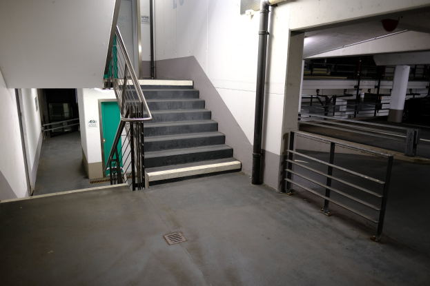 Leerer Parkhaus mit Treppe, die nach oben führt, Geländern auf beiden Seiten und Rohren an den Wänden; einige Gegenstände im Hintergrund sichtbar.