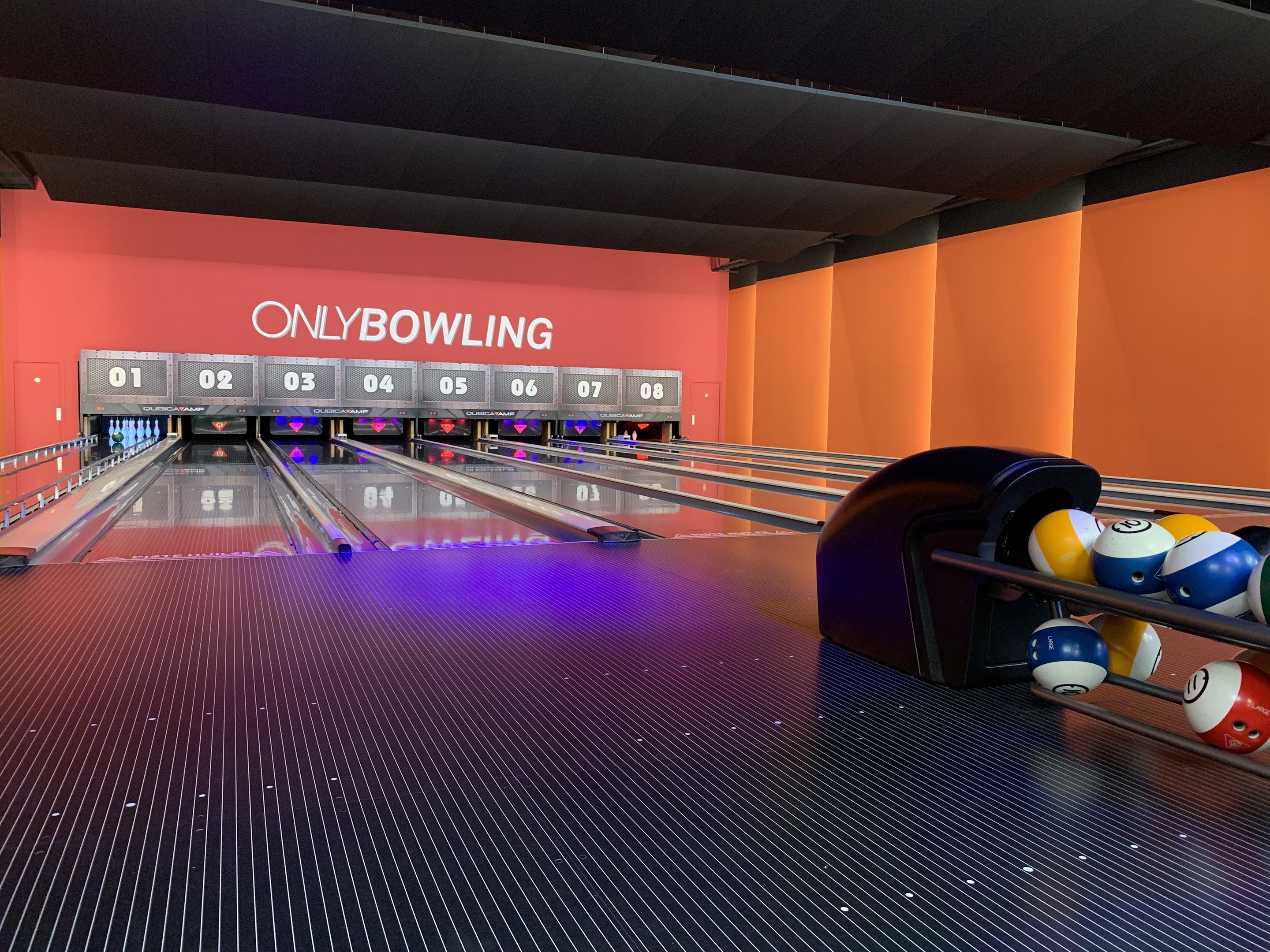 Eine Bowlingbahn mit einer Reihe von Bowlingkugeln auf der rechten Seite, aufgestellte Bowlingkegel in der Mitte und eine Wand mit Text im Hintergrund unter einer sichtbaren Decke.