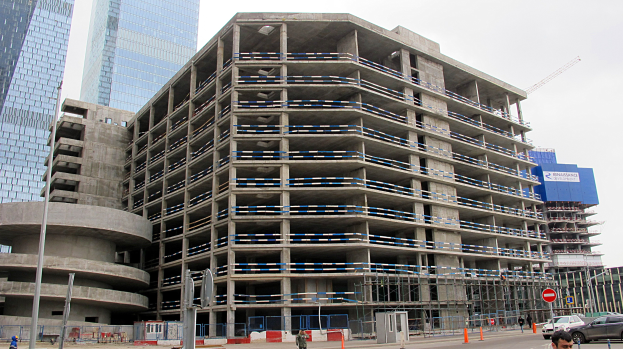 Ein großes Bauprojekt in einer städtischen Umgebung, umgeben von Wolkenkratzern, Fahrzeugen, Fußgängern, Verkehrsinfrastruktur und Baumaschinen unter einem klaren Himmel.