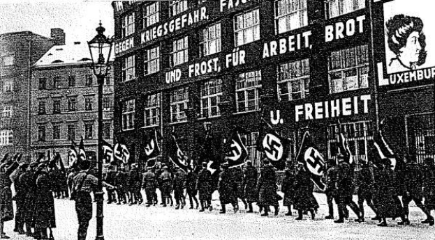 Schwarzes und weißes Foto einer Gruppe von Menschen, die mit Fahnen und Transparenten die Straße entlangmarschieren, mit einem Laternenmast links und einem Gebäude mit einer Tafel, auf der "Deutsche Nationalsozialistische Arbeiterpartei in Berlin" steht, rechts.