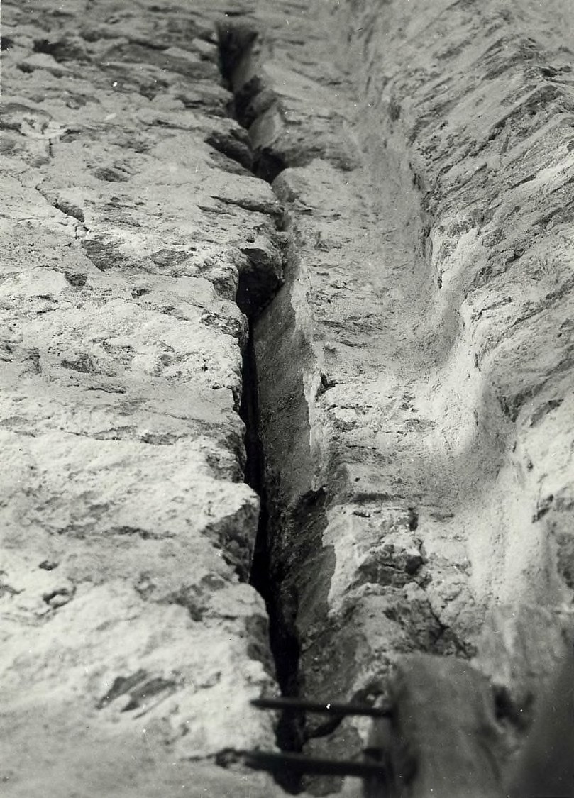 Schwarzes und weißes Foto eines großen Risses im Boden, wahrscheinlich ein Entwässerungskanal, mit einem sichtbaren Wasserzeichen auf der linken Seite.