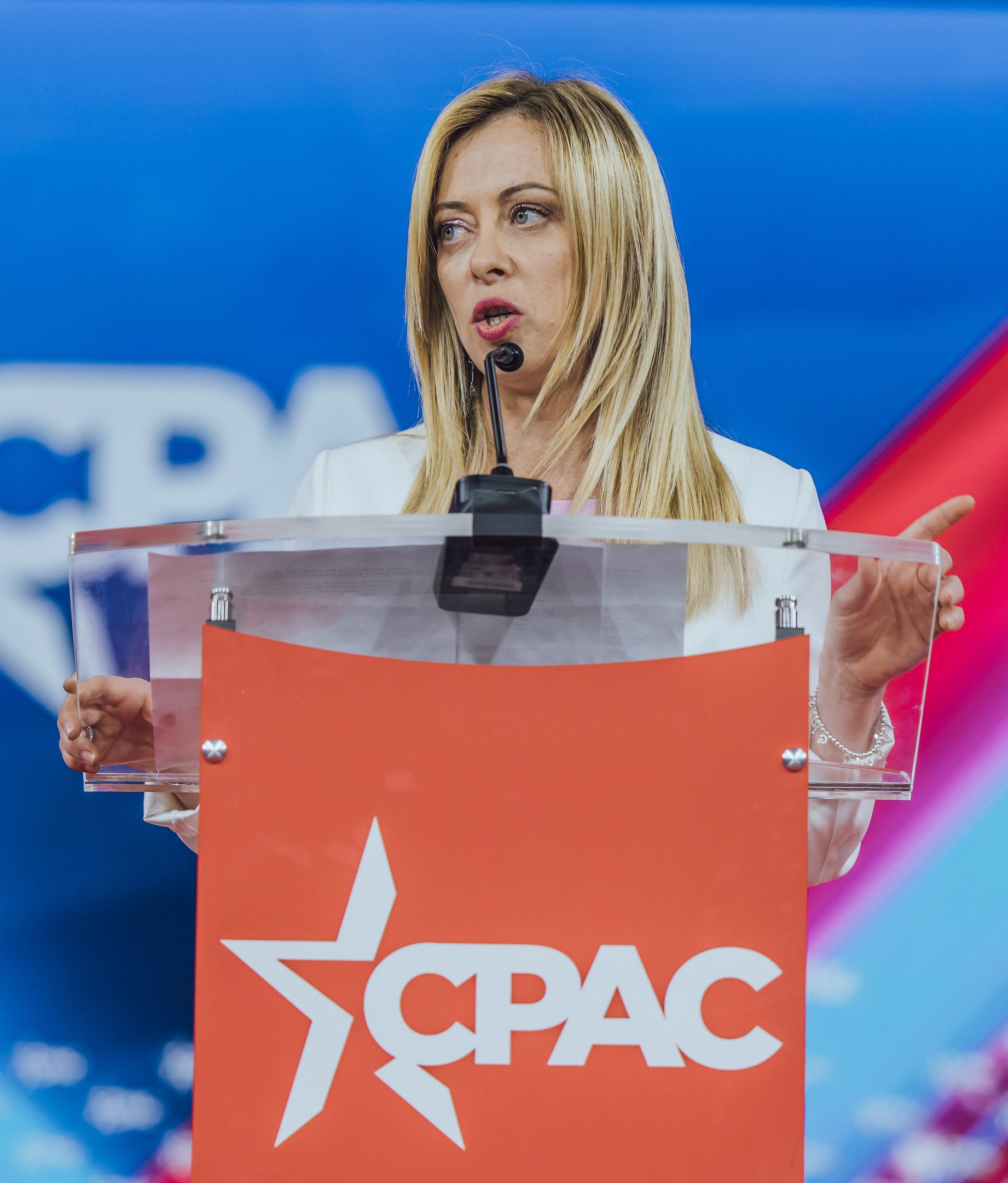 Frau in weißem Kleid, die an einem Rednerpult mit Mikrofon spricht, wahrscheinlich eine Politologin, vor einem unscharfen Hintergrund.