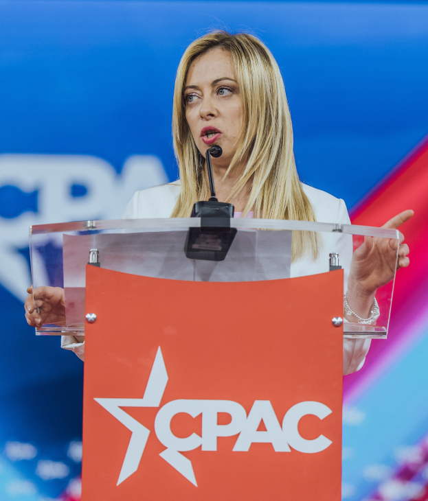 Frau in weißem Kleid, die an einem Rednerpult mit Mikrofon spricht, wahrscheinlich eine Politologin, vor einem unscharfen Hintergrund.