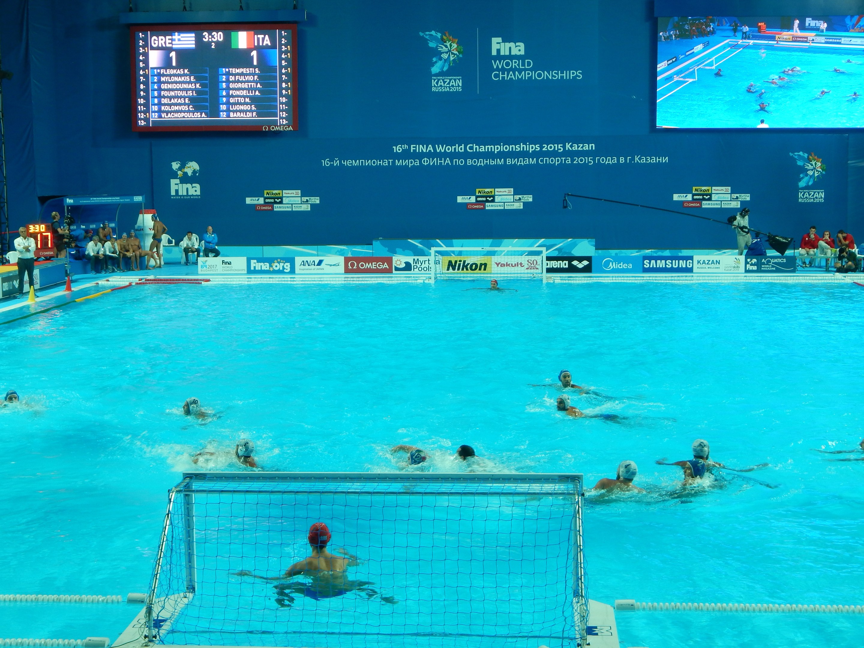 Wasserballspieler bei den 2016 FINA-Weltmeisterschaften in Kazan im Pool gegeneinander antretend, mit Zuschauern auf der linken Seite und einem Scoreboard mit Event-Branding im Hintergrund.