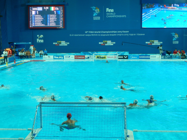 Wasserballspieler bei den 2016 FINA-Weltmeisterschaften in Kazan im Pool gegeneinander antretend, mit Zuschauern auf der linken Seite und einem Scoreboard mit Event-Branding im Hintergrund.