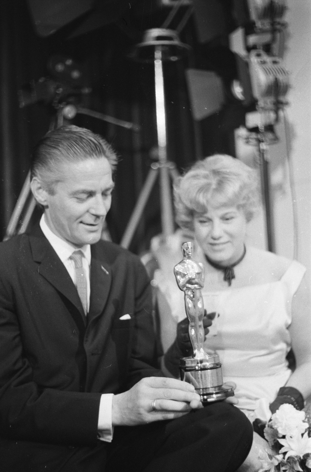 Ein Mann in einem schwarzen Blazer, weißem Hemd und Krawatte hält eine Oscar-Statue und sitzt neben einer Frau, die einen Blumenstrauß hält, beide in formeller Kleidung, mit Lichtern, die die schwarz-weiße Szene erhellen.