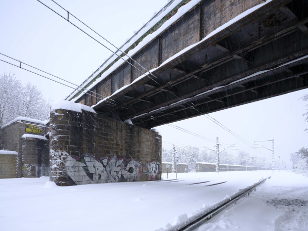 Eine schneebedeckte Bahnschiene unter einer Brücke mit Graffiti, Strommasten, Bäumen und einem Himmel im Hintergrund.