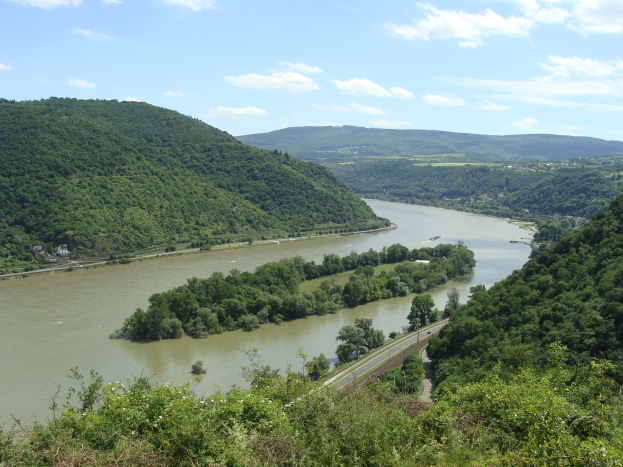 Panoramablick auf den Rhein, der sich durch eine grüne Landschaft schlängelt, mit einer Straße daneben und einem Himmel voller weißer Wolken.