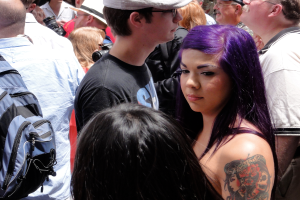 Eine Gruppe von Menschen, die stehen, mit einem Rucksack links, einer Frau mit einem Handtattoo rechts, einer Person in der Mitte mit einer Mütze und einer Kappe und einigen Individuen mit Kameras.