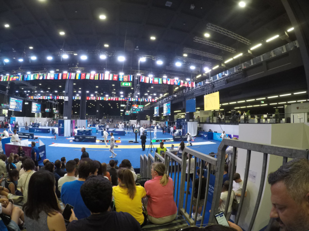 Eine große Menschenmenge auf Tribünen in einer Indoor-Sportarena, die einer Bühne gegenüberliegen, mit Stehplätzen auf dem Boden, sichtbaren Bildschirmen, Fahnen und Deckenleuchten.