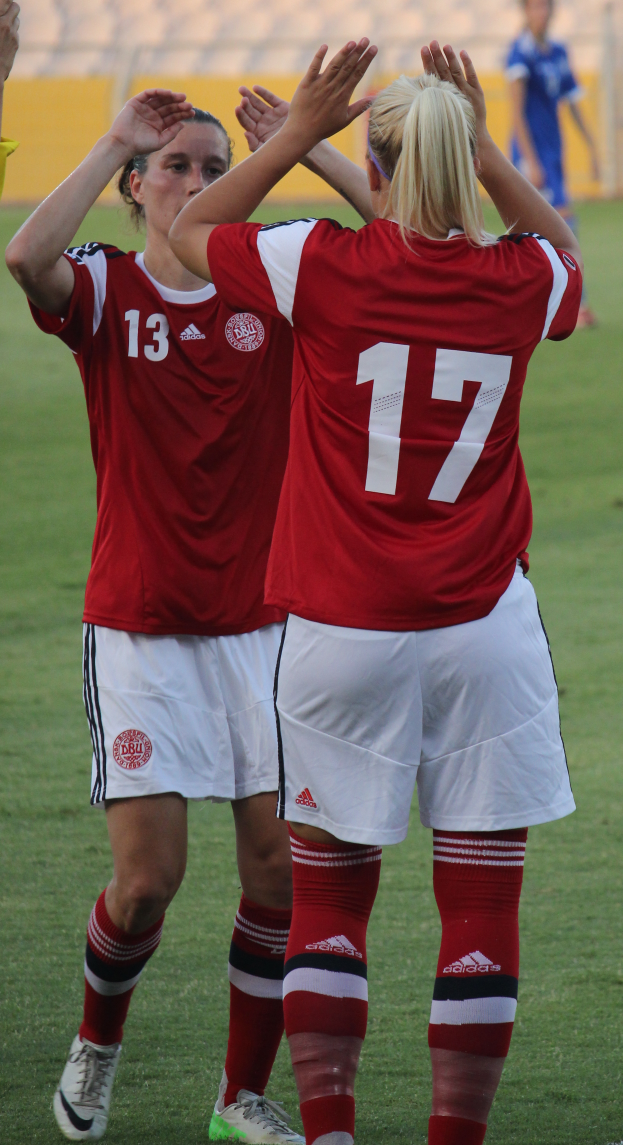 Zwei Frauen in roten T-Shirts und weißen Shorts stehen auf einem Fußballfeld und klatschen in die Hände, im Hintergrund eine Person in einem blauen Kleid, eine gelbe Tafel und einige Stühle.