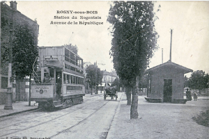 Ein Schwarz-Weiß-Foto einer Tram auf einer Straße in Rosny-sous-Bois, Frankreich, umgeben von Gebäuden, Bäumen, Pfählen und Menschen auf dem Gehweg, mit dem Himmel im Hintergrund und Text oben auf dem Bild.