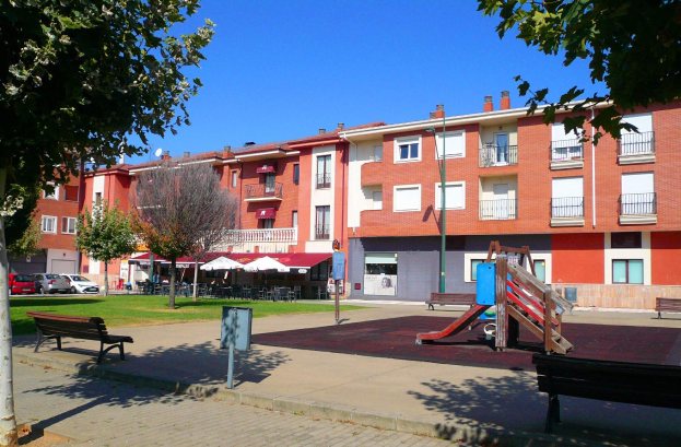 Ein Park mit einem Spielplatz, umgeben von Gebäuden mit Bänken, Bäumen, Gras, einem Pfahl, einem Schild, einer Straßenlaterne, Fahrzeugen, Zelten, Menschen unter einem Dach und einem bewölkten Himmel.