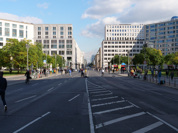 Eine belebte Straßenkreuzung in Berlin, Deutschland, mit Fußgängern und Radfahrern auf der Straße, hohen Gebäuden mit Fenstern, Bäumen, Laternen und Schildern unter einem bewölkten Himmel.
