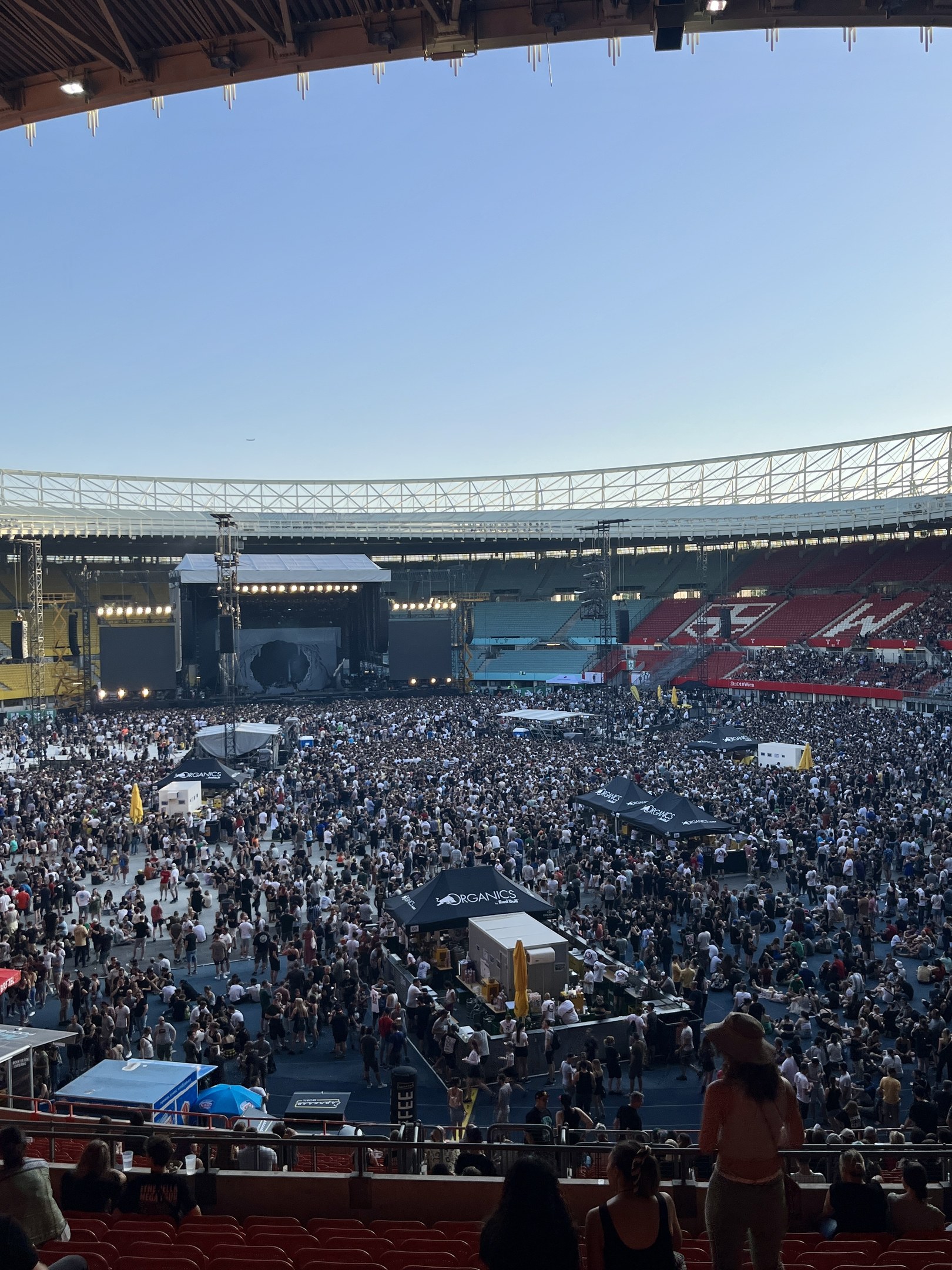 Große Menschenmenge auf Sitzplätzen und Stehplätzen in einem Stadion mit Zelten, Geländern und Überdachung unter einem Himmel als Hintergrund, was auf ein Konzert in der O2 Arena in London hindeutet.