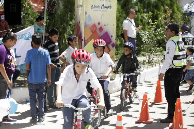 Kinder fahren mit Fahrrädern eine Straße entlang, die von Verkehrskegeln gesäumt ist, einige tragen Helme, andere stehen in der Nähe, mit einer Fahne, Bäumen und Gebäuden im Hintergrund.