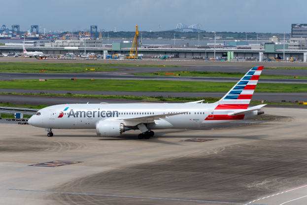 Ein American-Airlines-Flugzeug auf der Rollbahn eines Flughafens, umgeben von Gras, Fahrzeugen, Gebäuden, Polen, Kränen und anderen Gegenständen, mit Bäumen und einem klaren blauen Himmel im Hintergrund.