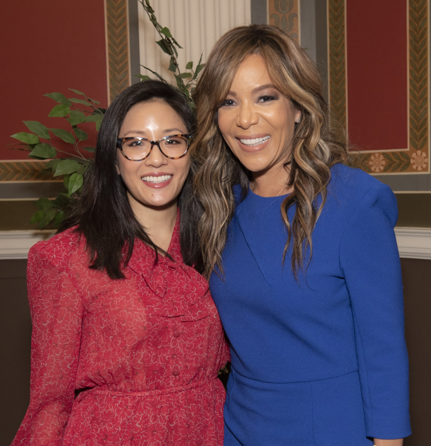 Zwei Frauen stehen nebeneinander und lächeln vor einer Wand mit einer Pflanze und einem Vorhang auf der 2019 Women in Film Pre-Oscar-Lunch; die Frau rechts trägt ein blaues Kleid, die Frau links trägt ein rotes Kleid und eine Brille.