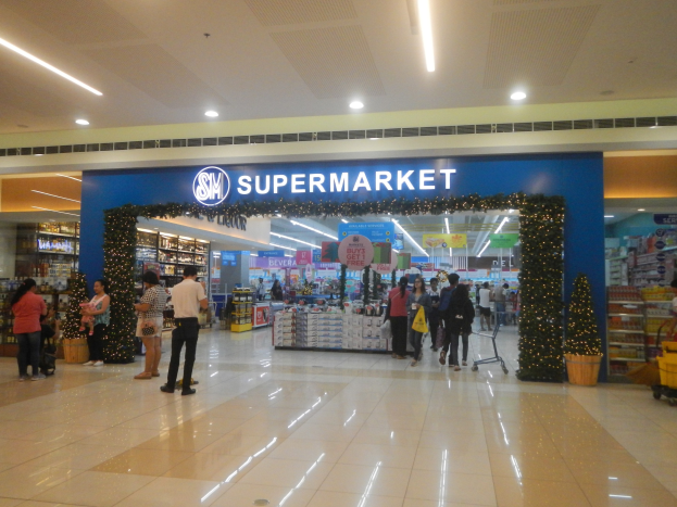 Eine Gruppe von Menschen, die vor einem Supermarkt stehen, einige halten Einkaufstaschen, mit einem Einkaufswagen, Topfpflanze, Regalen, Schildern und Deckenleuchten im Hintergrund.