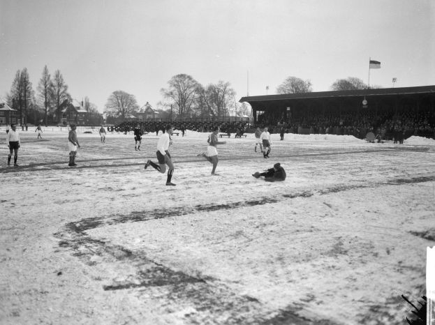 Schwarzes und weißes Bild von Menschen, die Fussball auf einem schneebedeckten Feld mit Bäumen, Häusern, Pfählen, Fahnen und einer Hütte im Hintergrund spielen.
