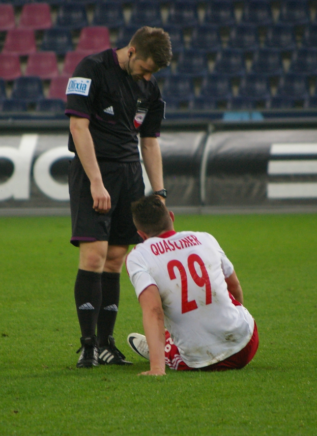Ein Fussballspieler sitzt auf dem Boden neben einem Schiedsrichter in einem Stadion, beide tragen Sportkleidung, mit Tafeln und Stühlen im Hintergrund.