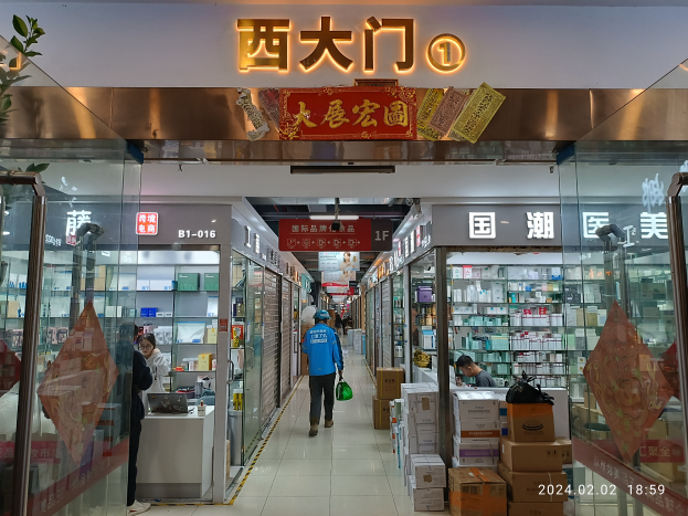 Das Innere einer chinesischen Apotheke mit Menschen, die hindurchgehen, Glaswänden auf beiden Seiten, einer Pflanze auf der linken Seite, Regalen, die mit verschiedenen Gegenständen gefüllt sind, Kartonagen und anderen Gegenständen, die auf dem Boden verstreut sind, und einem Namensschild mit Text oben.