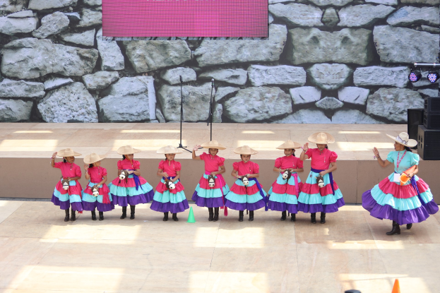 Gruppe junger Mädchen in bunten Kleidern und Hüten auf einer Bühne mit Verkehrskegeln, Mikrofonen, Lautsprechern, Scheinwerfern und einem Bildschirm mit Text im Hintergrund, während das Publikum von unten zusieht.