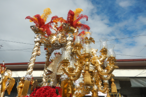 Ein prunkvoller gold-roter Festwagen mit Blumen und anderen Dekorationen bei einem Karnevalsumzug, im Hintergrund ein Gebäude, Strommasten mit Kabeln und ein bewölkter Himmel.