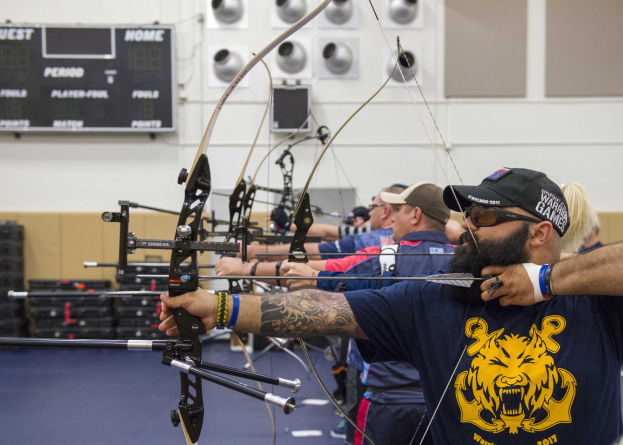 Gruppe von Menschen in Mützen beim Bogenschießen in der Halle während der World Archery Championships 2017, mit einer Texttafel links und Gegenständen auf dem Boden rechts.