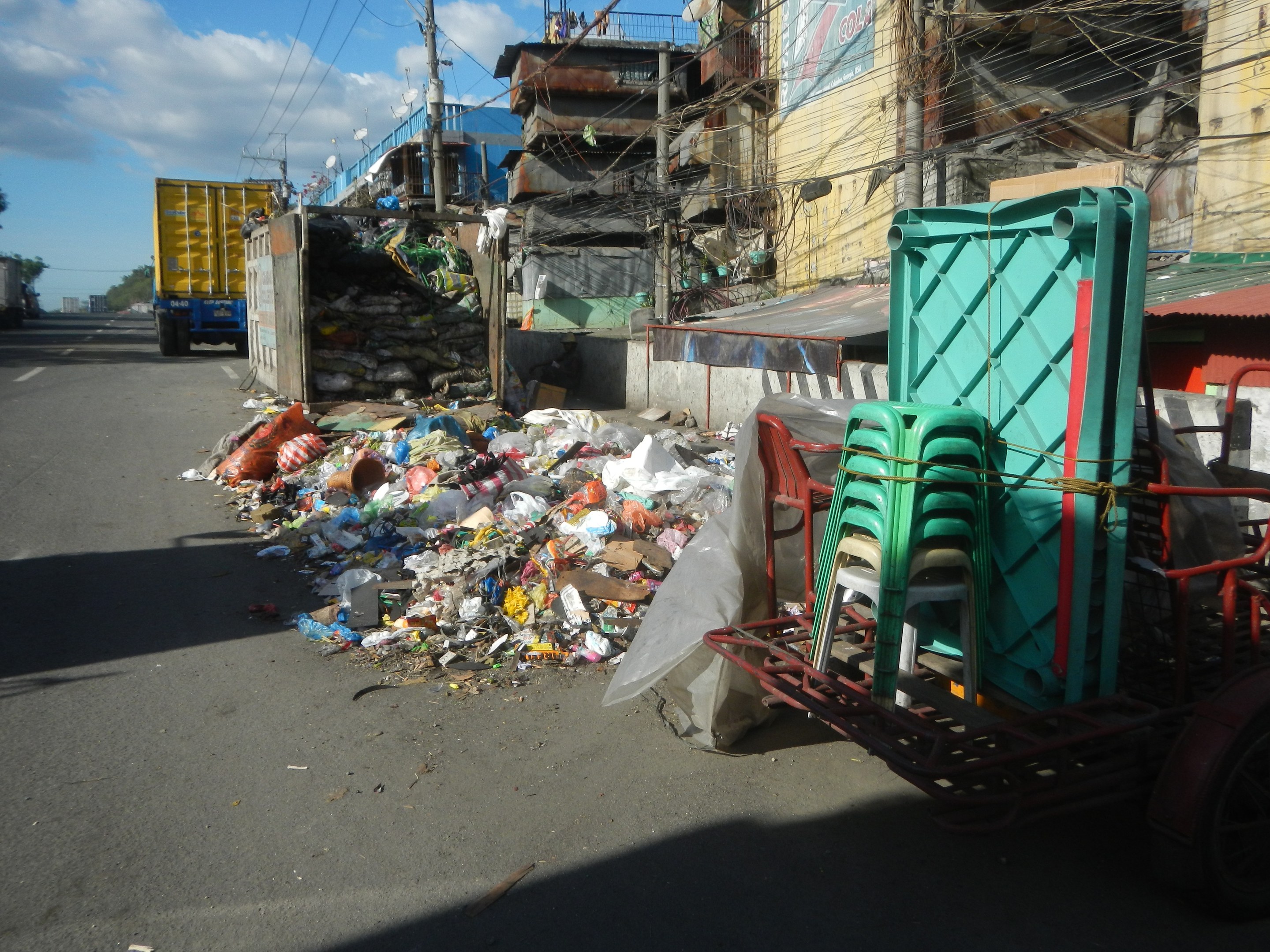 Ein Lastwagen neben einem Müllhaufen auf einer Straße geparkt, mit einem Wagen rechts daneben, der mit Plastikstühlen beladen ist, und Gebäuden, Strommasten, Bäumen und einem bewölkten Himmel im Hintergrund.