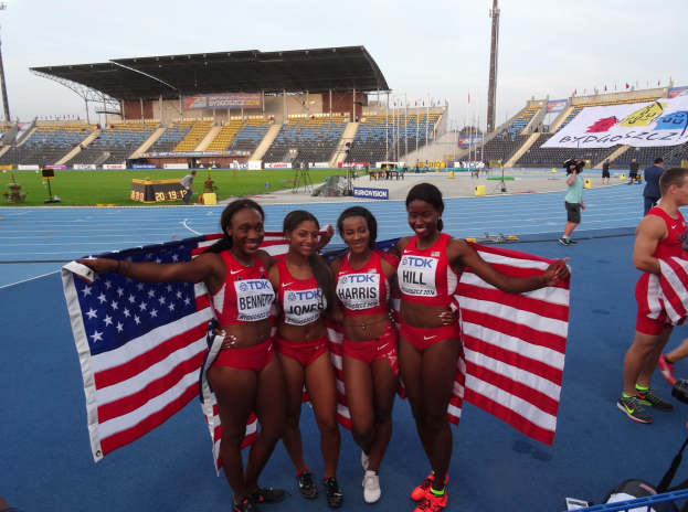 Vier Frauen in Sportkleidung stehen zusammen auf einer Laufbahn, lächeln und halten amerikanische Flaggen, mit einem Stadion und Zuschauern im Hintergrund.