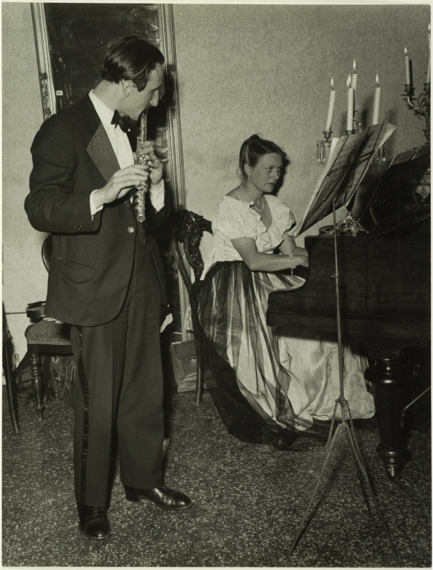 Ein Mann im Smoking spielt Klarinette, während er neben einer Frau steht, die an einem Piano sitzt und Noten auf dem Ständer hat, und ein gerahmtes Bild ist im Hintergrund an der Wand zu sehen.