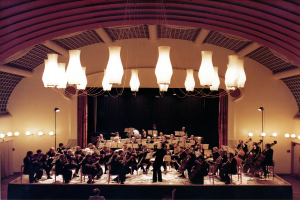 Eine Gruppe von Menschen, die Geigen spielen, alle in Schwarz gekleidet, mit einem Mann unten und Lichtern, die von der Decke hängen, vor einem Hintergrund aus einer Wand mit einem Vorhang.