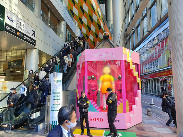 Eine Gruppe von Menschen steht vor einem Gebäude mit Glaswänden, Säulen und Texttafeln, einige tragen Masken, während eine Rolltreppe mit Menschen zu sehen ist; ein pinker Kasten in der Mitte zeigt Schaufensterpuppen und trägt den Text "Netflix' neue Netflix-Bühne in Tokio."
