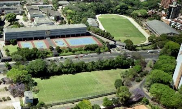 Luftbild eines Fußballfeldes auf dem Campus der Universität Sydney, umgeben von Bäumen, Gebäuden und Fahrzeugen, mit einem neuen Sportkomplex in Planung.